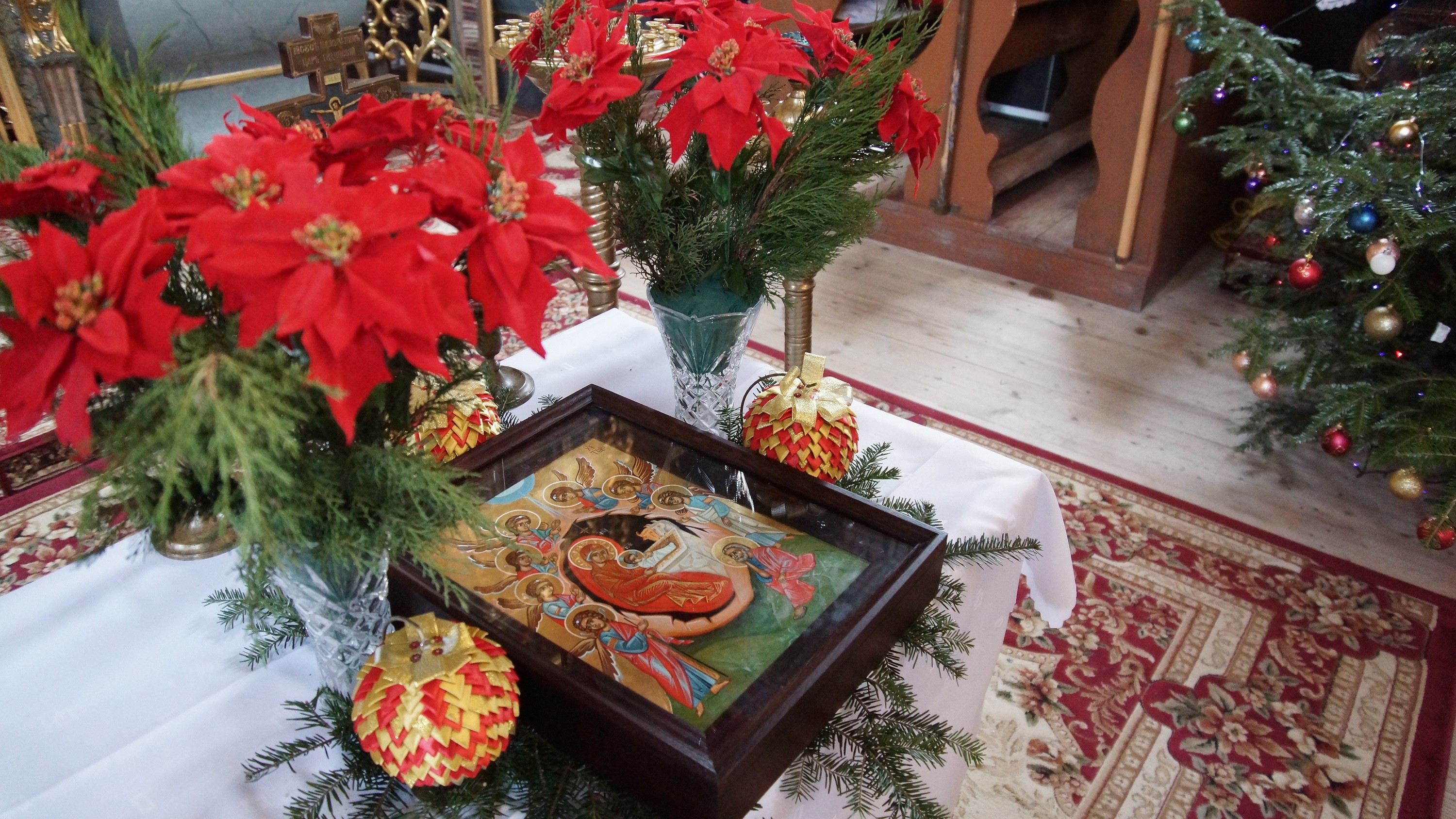 ikona z przedstawieniem Bożego Narodzenia. Obraz za szkłem, w grubej drewnianej ramie leży na niewielkim stoliku, nakrytym białym obrusem. Obok ikony wysokie szklane wazony z czerwonymi kwiatami.