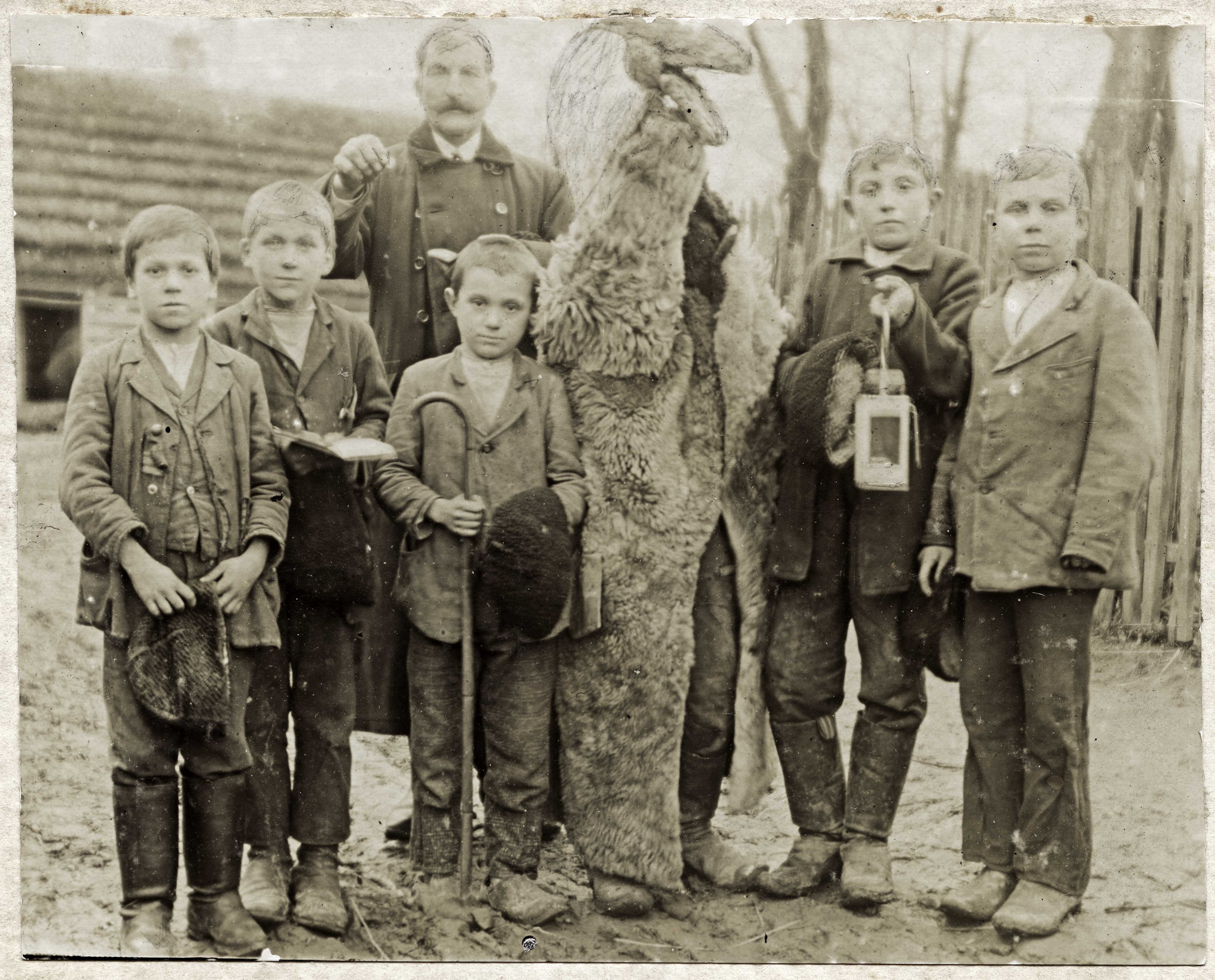 czarnobiałe zdjęcie - grupka chłopców pozuje do zdjęcia, wśród nich postać przebrana za kozę oraz odświętnie ubrany mężczyzna